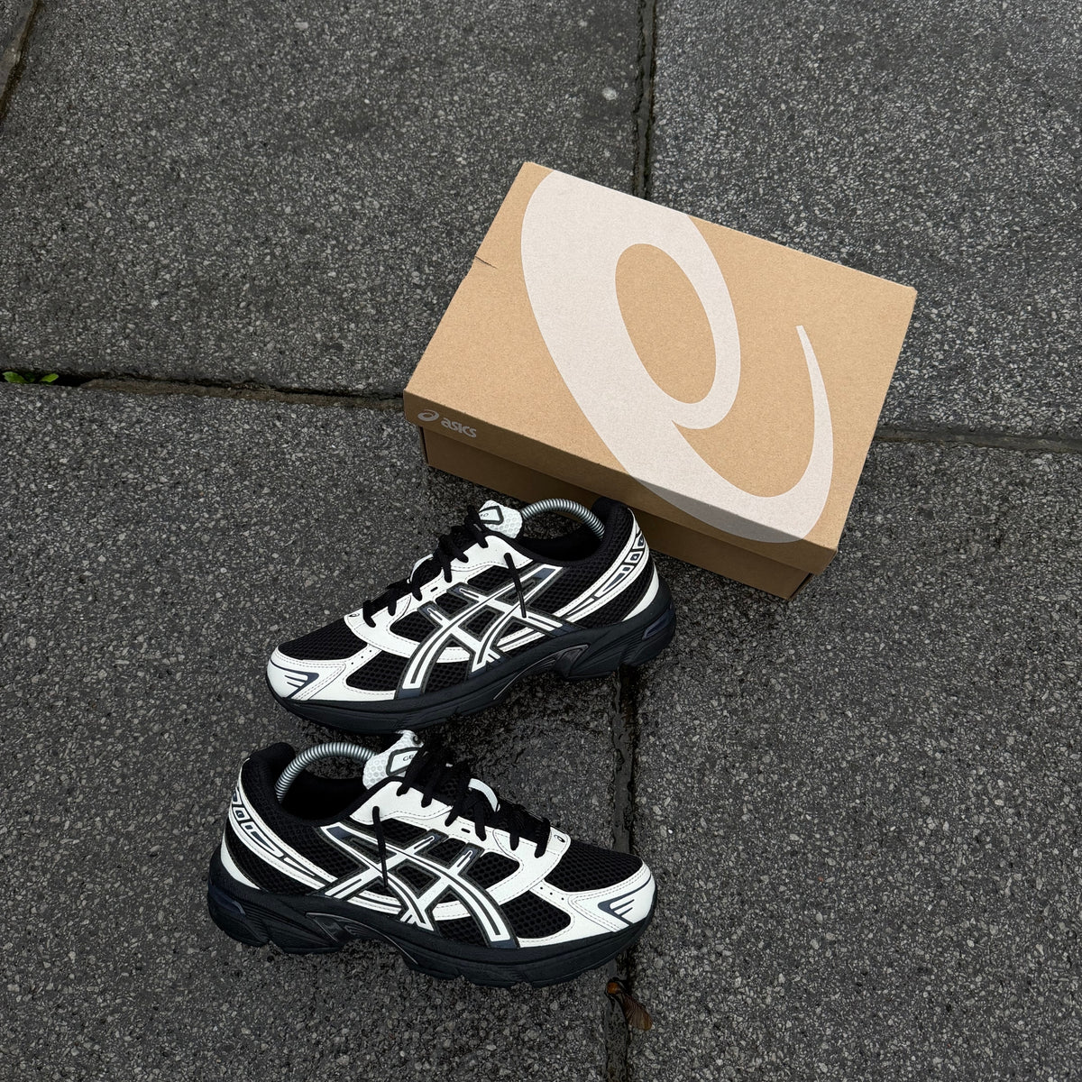Pair of ASICS GEL-1130 Black Cream trainers photographed on grey pavement beside original ASICS shoebox