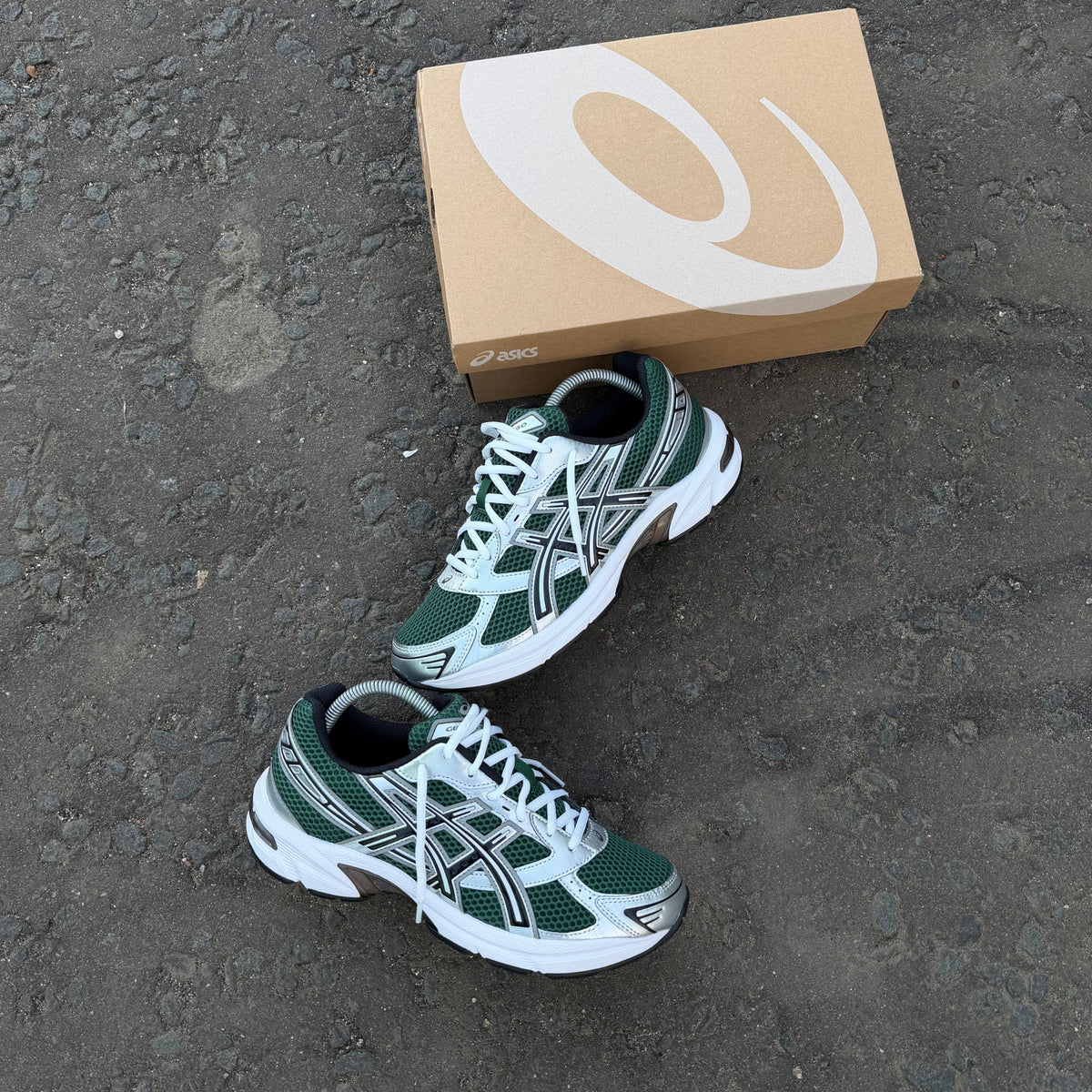 Pair of ASICS GEL-1130 Evergreen Black trainers displayed on dark pavement beside original ASICS shoebox