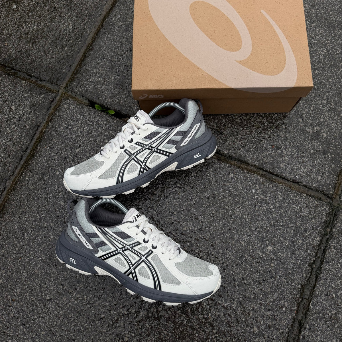 Pair of ASICS Gel-Venture 6 Olive Grey Cream trainers displayed on grey pavement beside an ASICS shoebox