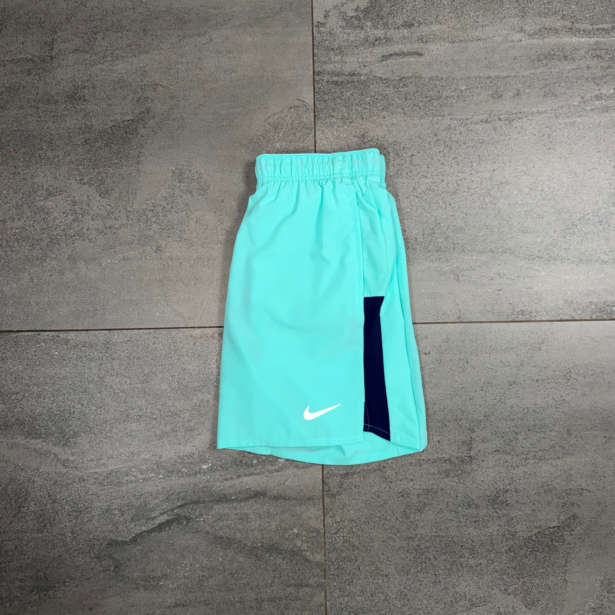 A pair of Nike athletic shorts in a bright mint green color, laid flat on a gray tiled floor. The shorts feature a white Nike swoosh logo near the hem and a dark navy blue vertical panel on the side.