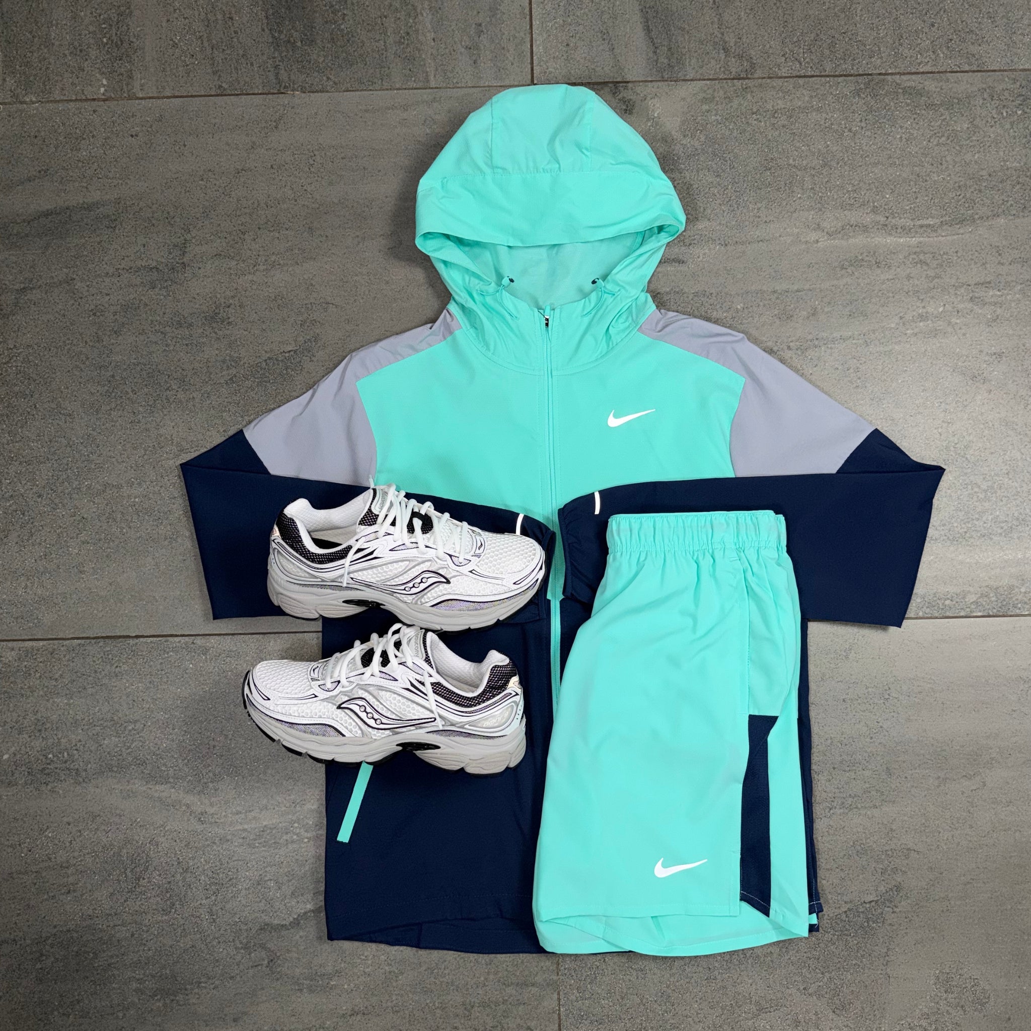 A Nike athletic outfit laid flat on a gray tiled floor, featuring a mint green and gray zip-up hooded jacket, navy shorts, and white sneakers.