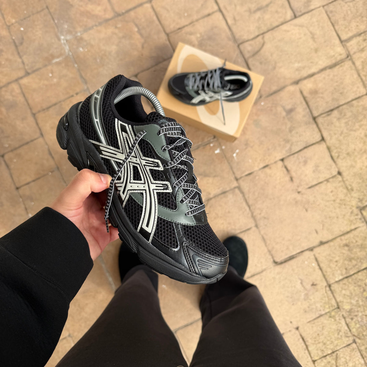 ASICS Gel-1130 Black Glacier Grey trainer held outdoors with the matching pair displayed behind