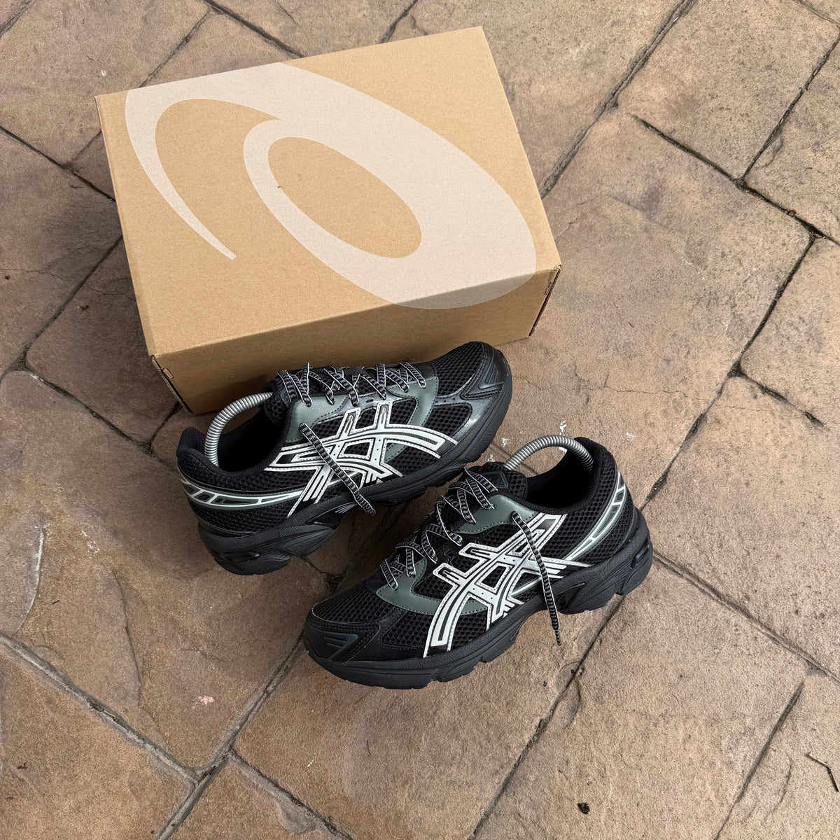Pair of ASICS Gel-1130 Black Glacier Grey trainers displayed outdoors beside their ASICS shoe box