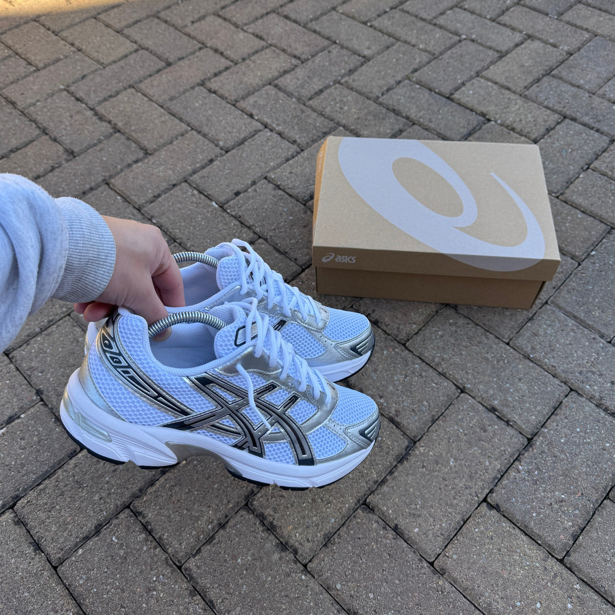 Pair of ASICS Gel-1130 White Pure Silver trainers displayed beside an ASICS shoe box outdoors